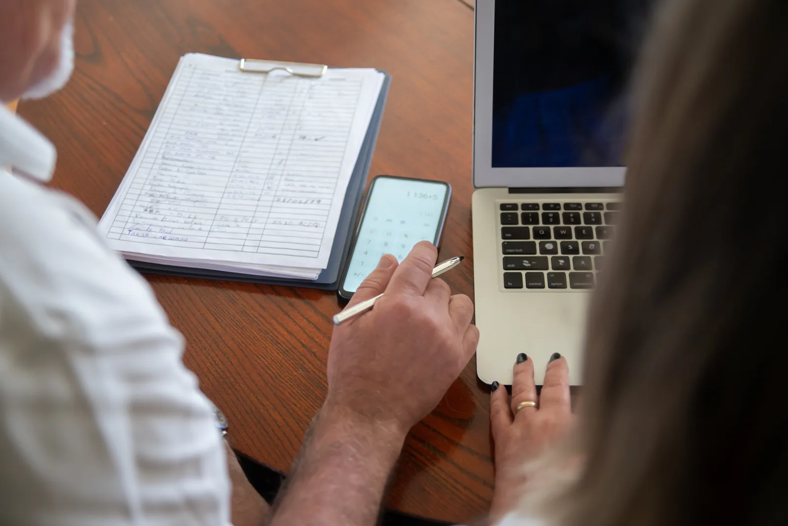 Two adults working at a desk with a laptop, smartphone, and documents. Collaborative workspace scene.