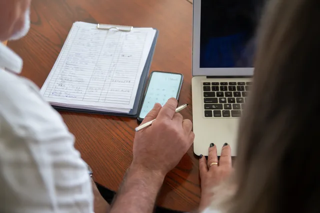Two adults working at a desk with a laptop, smartphone, and documents. Collaborative workspace scene. Survey tools for UK professional services marketing: free, paid and data protection questions